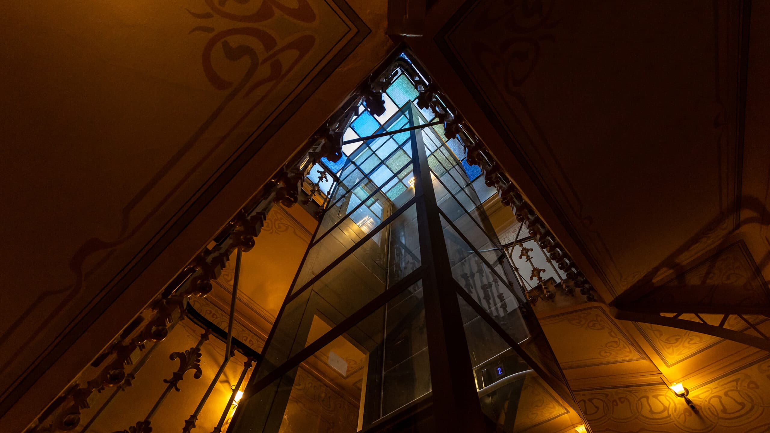 Bronze coloured Cibes A5000 platform lift in an old staircase, with multi-coloured glass ceiling. Photographed from below.