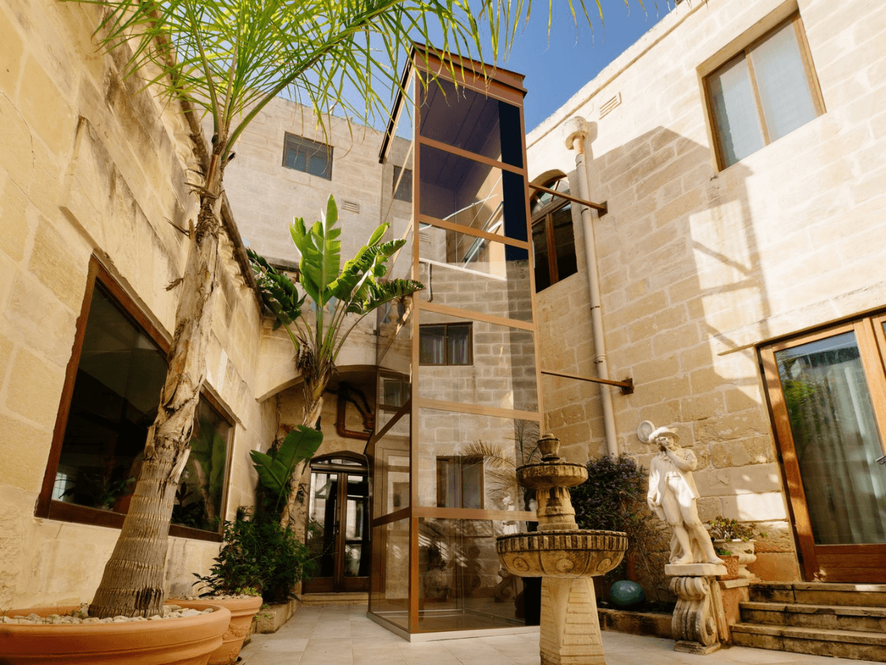 A stone courtyard with tall beige walls featuring large windows, a glass elevator, potted plants including a palm tree, a decorative fountain, and a statue of a man wearing a hat and holding a staff. home abroad in Croatia, home of people who have planned to retire in a foreign country.