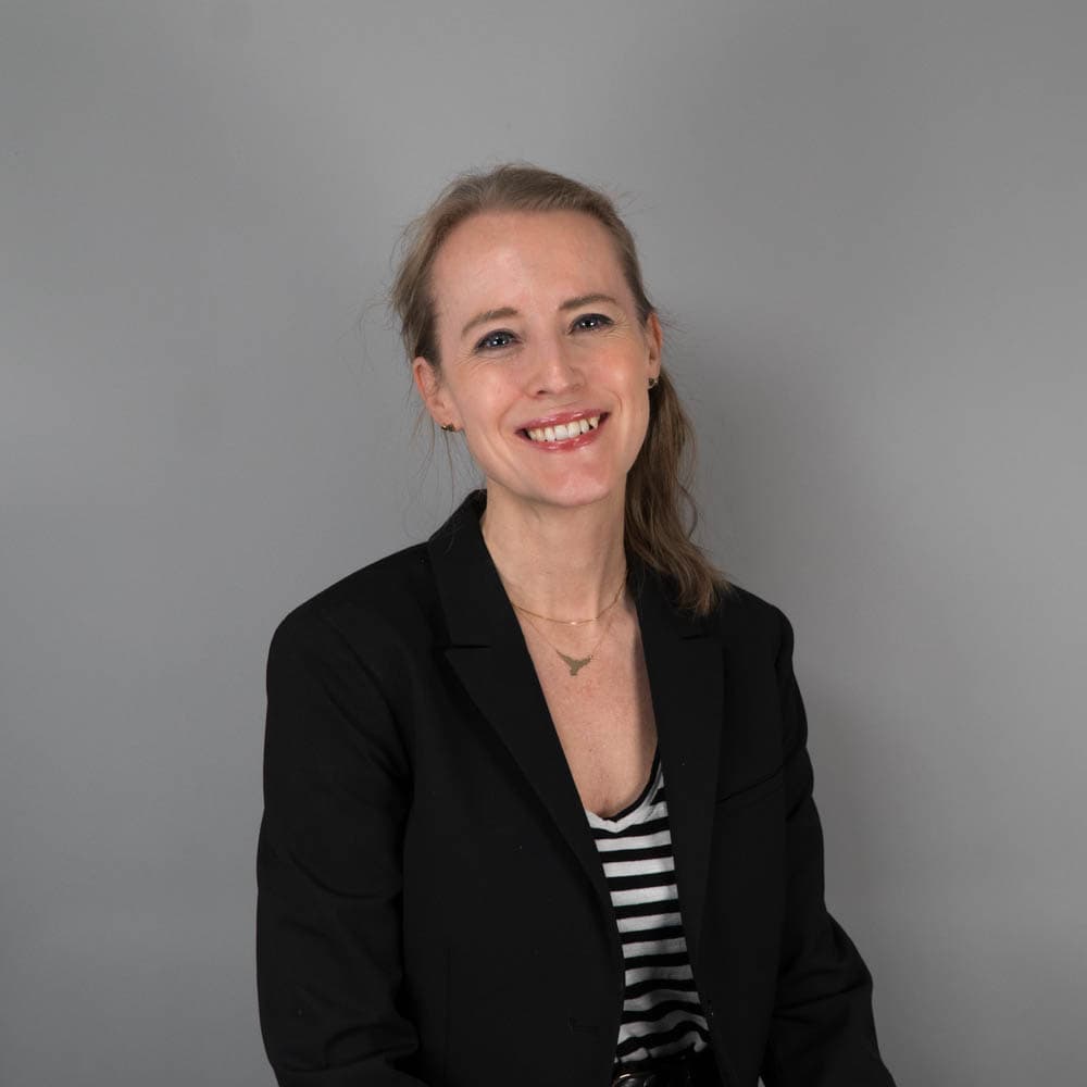 A woman with dark blonde hair tied back is smiling and wearing a black blazer over a black-and-white striped top, set against a plain gray background.