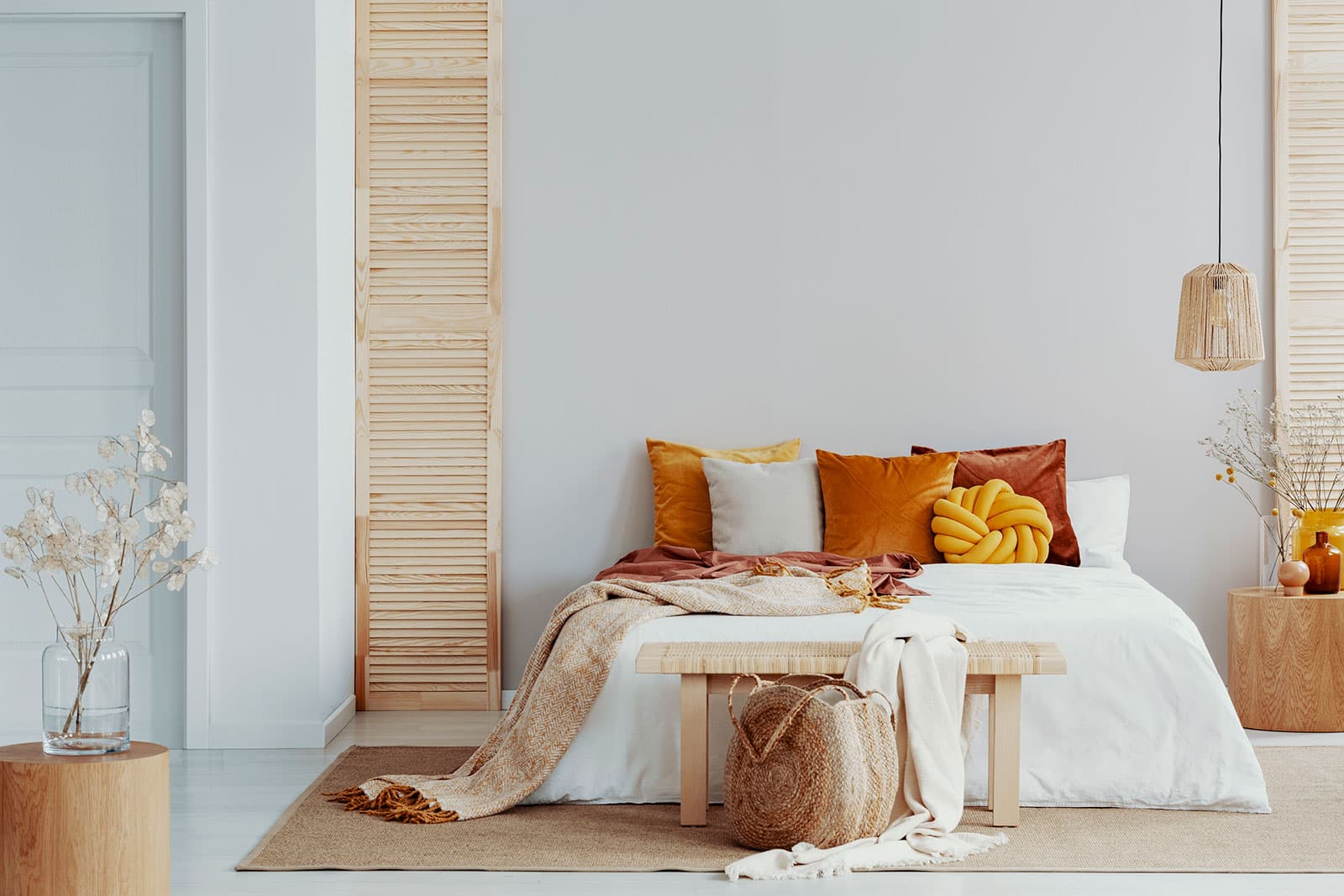Brown and orange pillows on white bed in natural bedroom interior with wicker lamp and wooden bedside table with vase