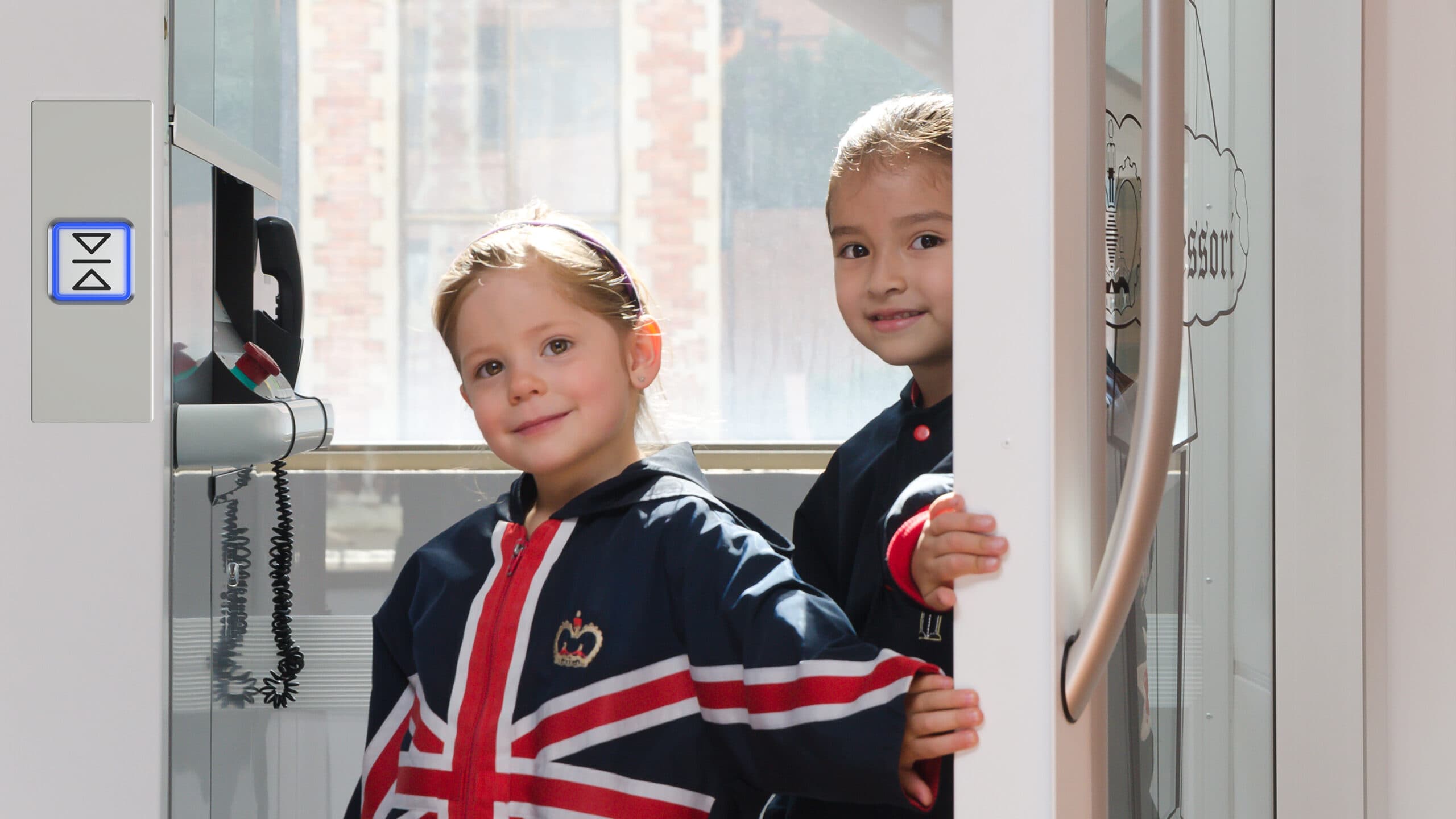 Two small school girls in blue sweatshirts with British flag motive in a white lift with open door.