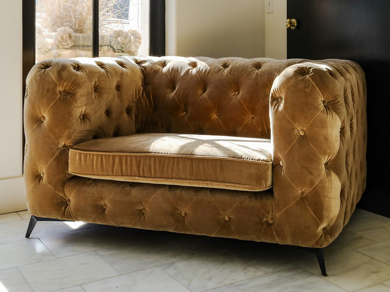 Brown, modern interpretation of the Chesterfield sofa design in cinnamon brown velvet, next to a window with black frames.