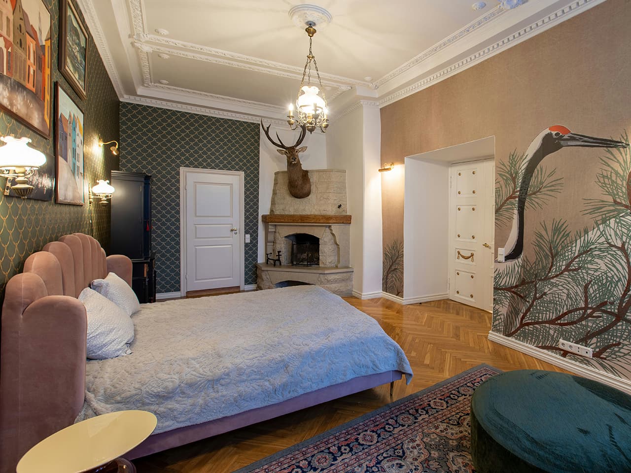 Maximalist style bedroom in turn of the century apartment, with art deco inspired bed, dusty pink wallpaper with cranes and patterned dark green wall paper and rustic fire place with a stuffed deer head.