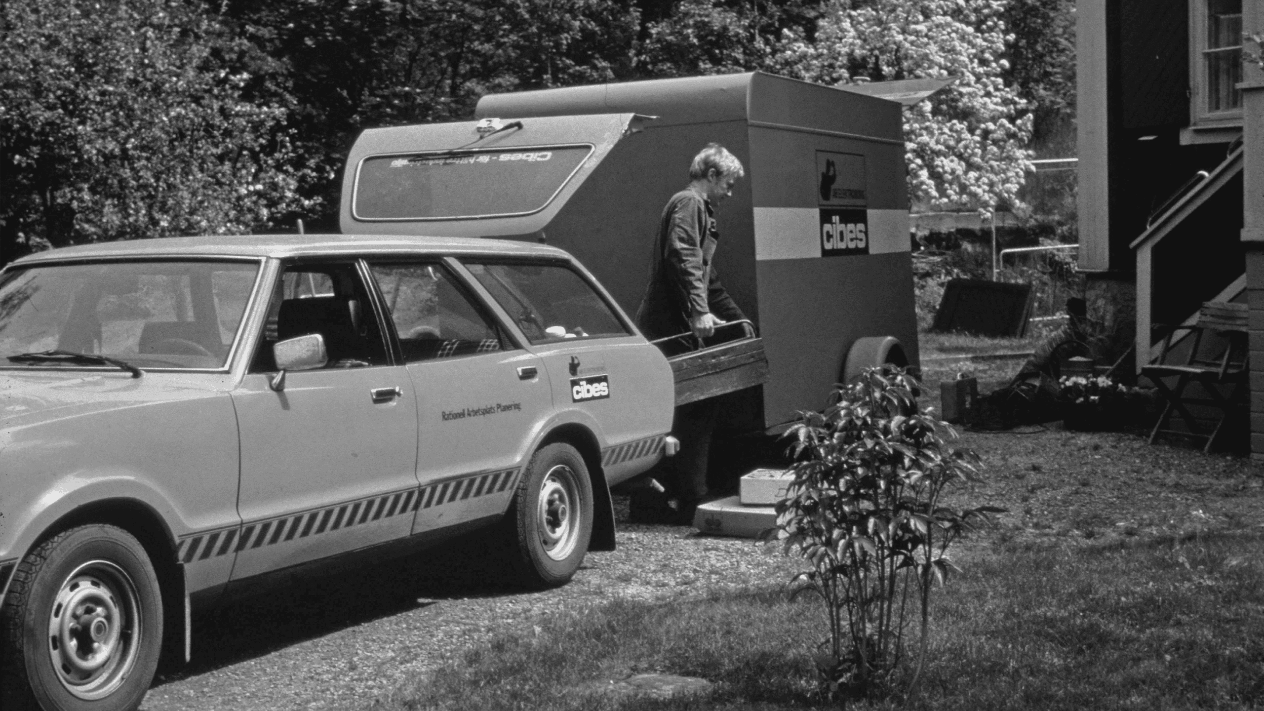 Old photograph in balck and white. A man stands near a trailer labelled “Cibes” attached to a station wagon with “Rational Architects Planning” written on its side. The scene shows a grassy area, a house, and blooming trees in the background.