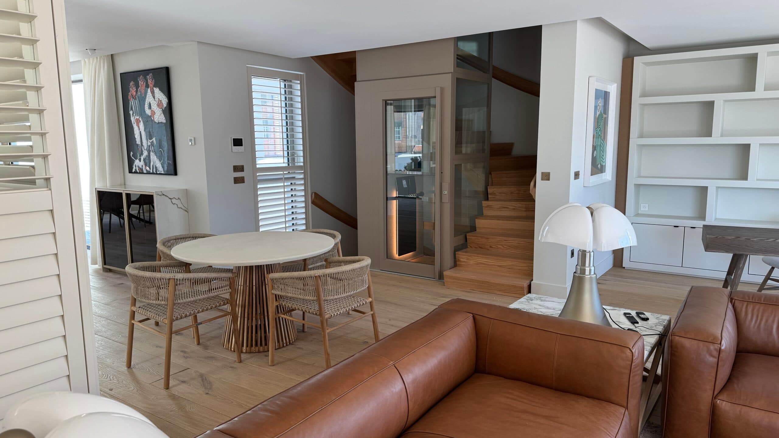 A modern living space featuring a small dining table with four woven chairs, a leather sofa, a white mushroom-shaped lamp, and wooden stairs leading to an upper level. The room includes a built-in elevator, white shelving, and a painting on the wall.