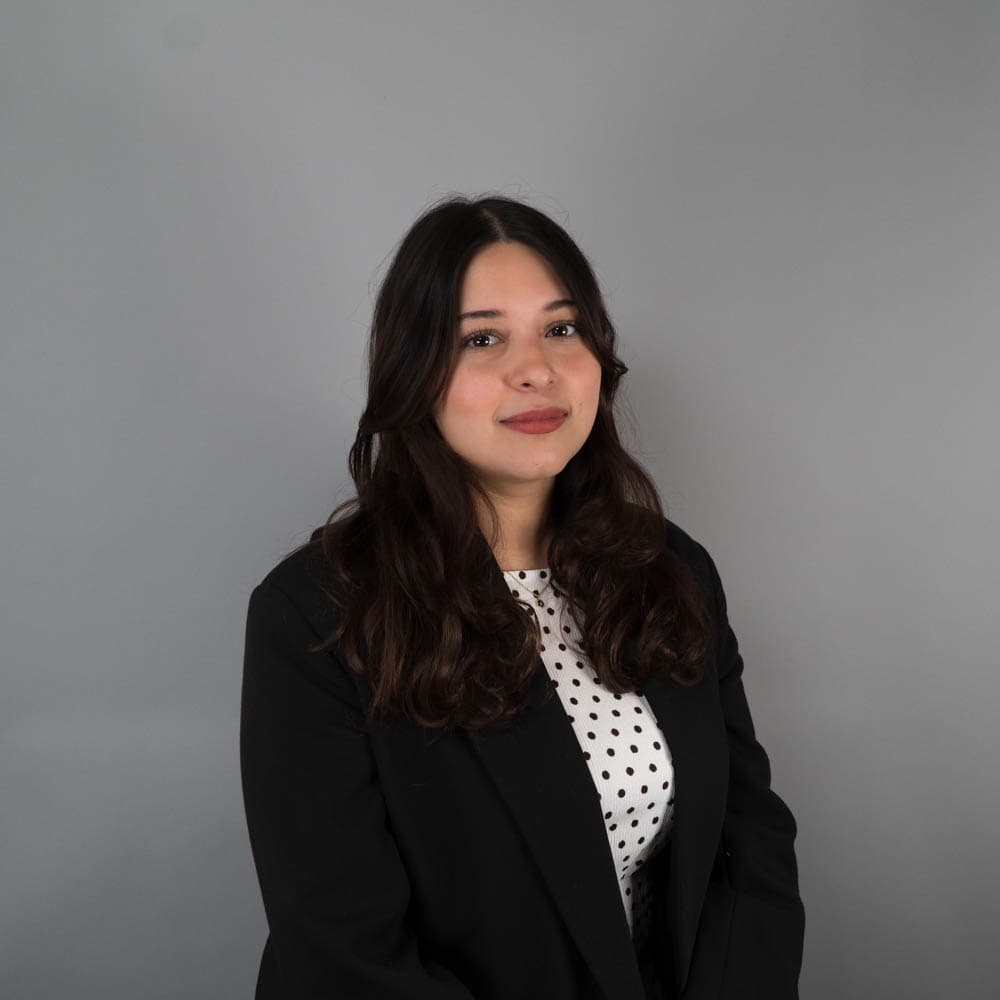 A woman with long dark hair sits against a plain gray background, wearing a black blazer and a white polka-dotted top.