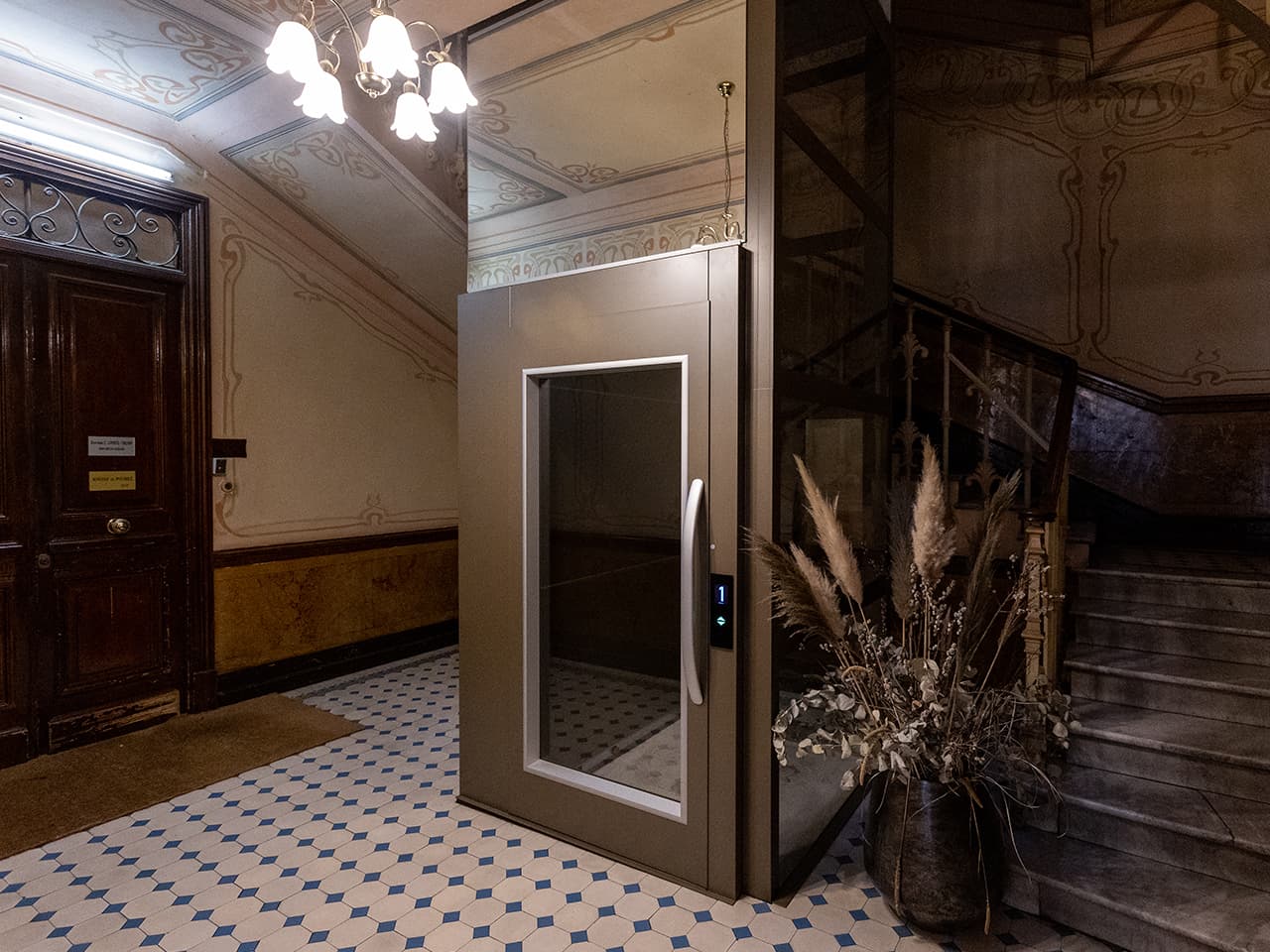 A modern enclosed elevator with a glass door and control panel displaying "1," situated in an old Art Nouveau stairwell with patterned floor tiles, ornate wall designs, a wooden door, and a staircase with dried plants in a decorative vase nearby.