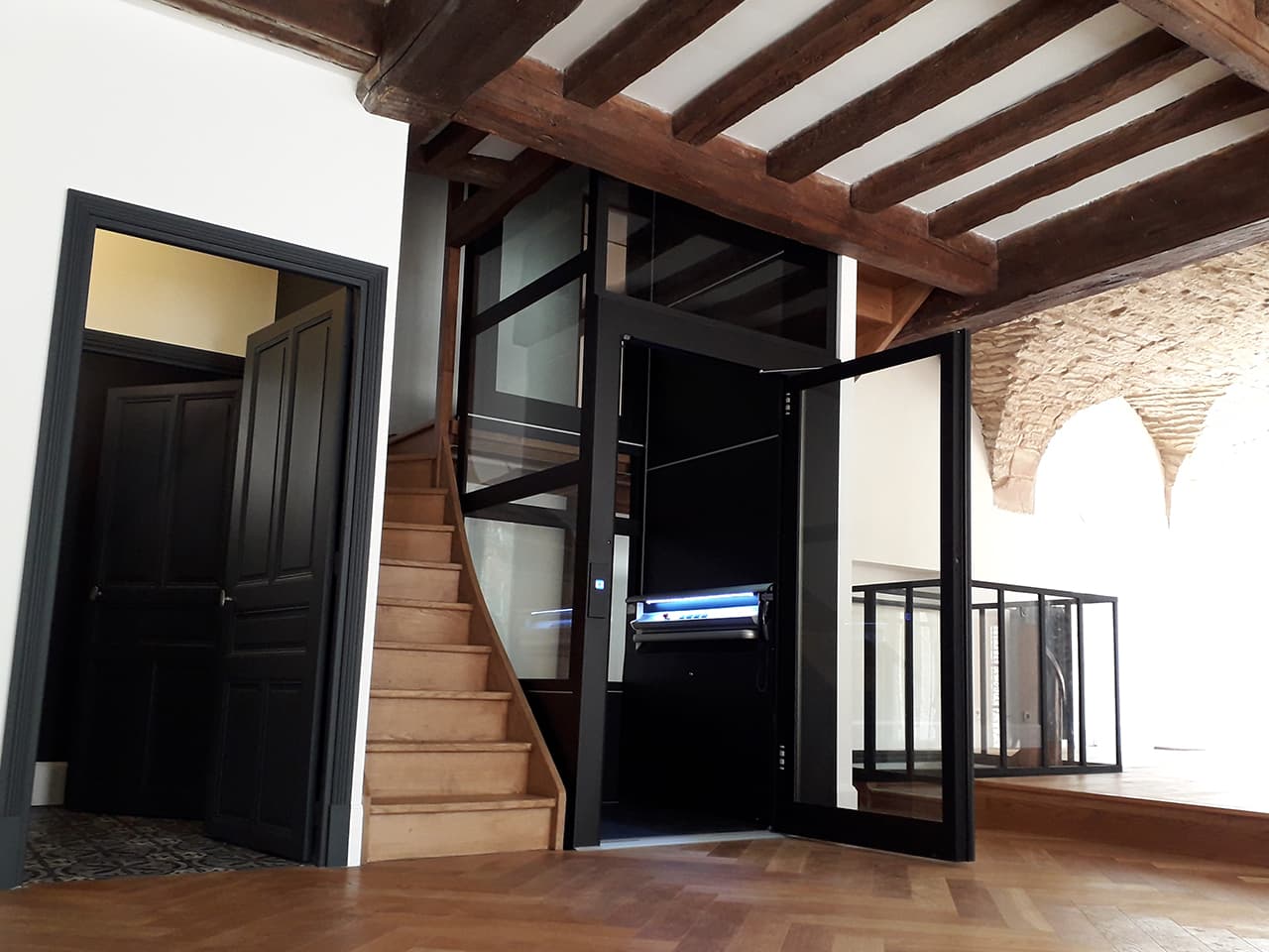 Wooden stairs of an old property next to a modern glass elevator with an open black-framed door, surrounded by exposed wooden beams and stone archways. A nearby doorway features a black ornated door leaf.