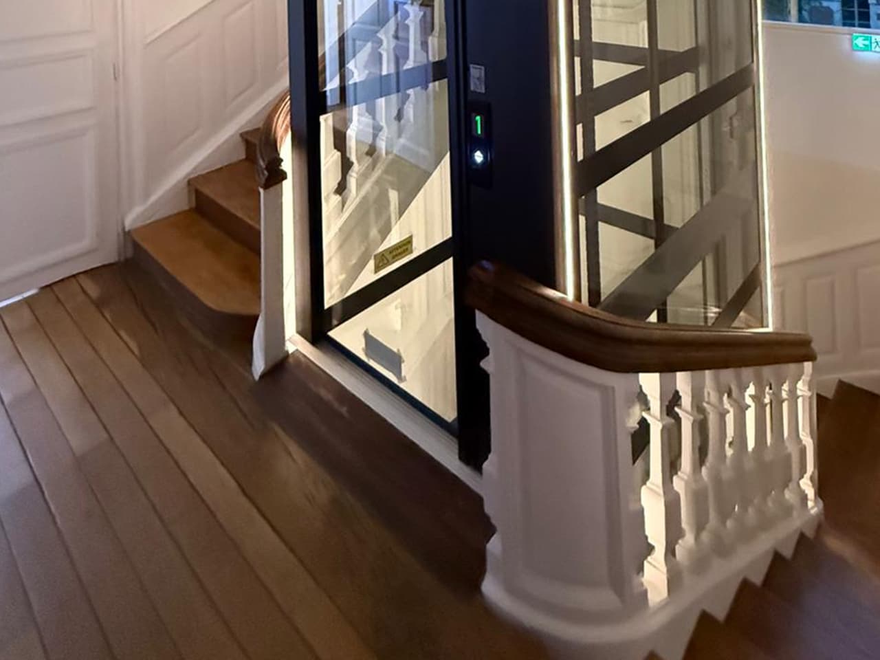 A modern glass elevator with illuminated edges is installed in a period wooden staircase with a white balustrade in a well-lit interior space featuring wooden floors and paneled walls.