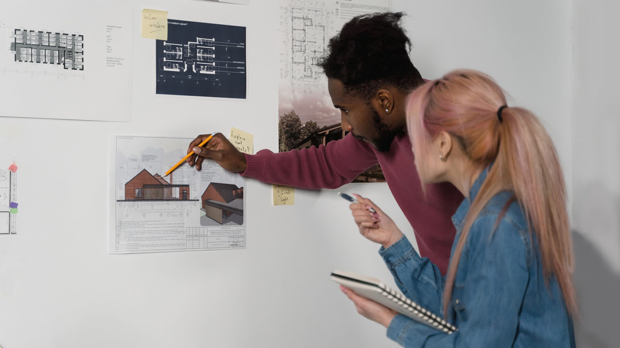 Two individuals, one male and one female figure, collaborate on architectural plans pinned to a white wall; one points to a house design with a pencil while the other holds a notebook and pen.