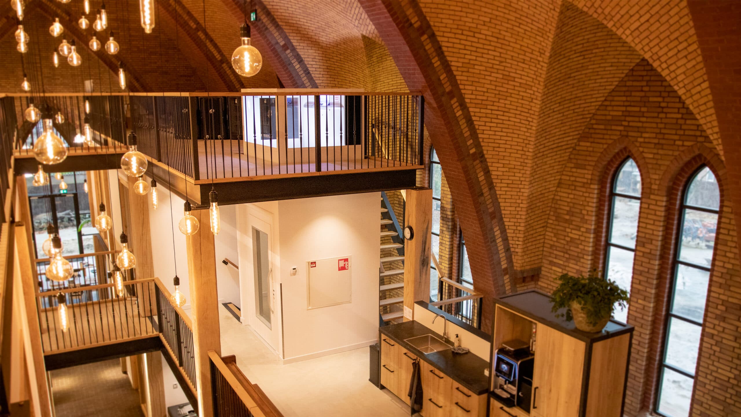 Interior view of a renovated old church with exposed brick arches, modern lighting fixtures, and a wooden kitchen area. Large windows let in natural light, and a staircase leads to a mezzanine level. The mezzanine level is equipped with a white Cibes A5000 platform lift with a gate for the top floor.