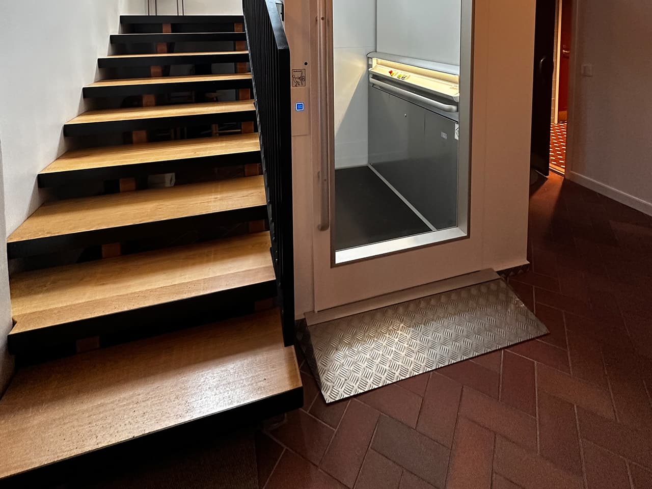 A wooden staircase with black railings is next to a white Cibes platform elevator with a metal access ramp leading to its entrance, all situated on a red tiled floor.