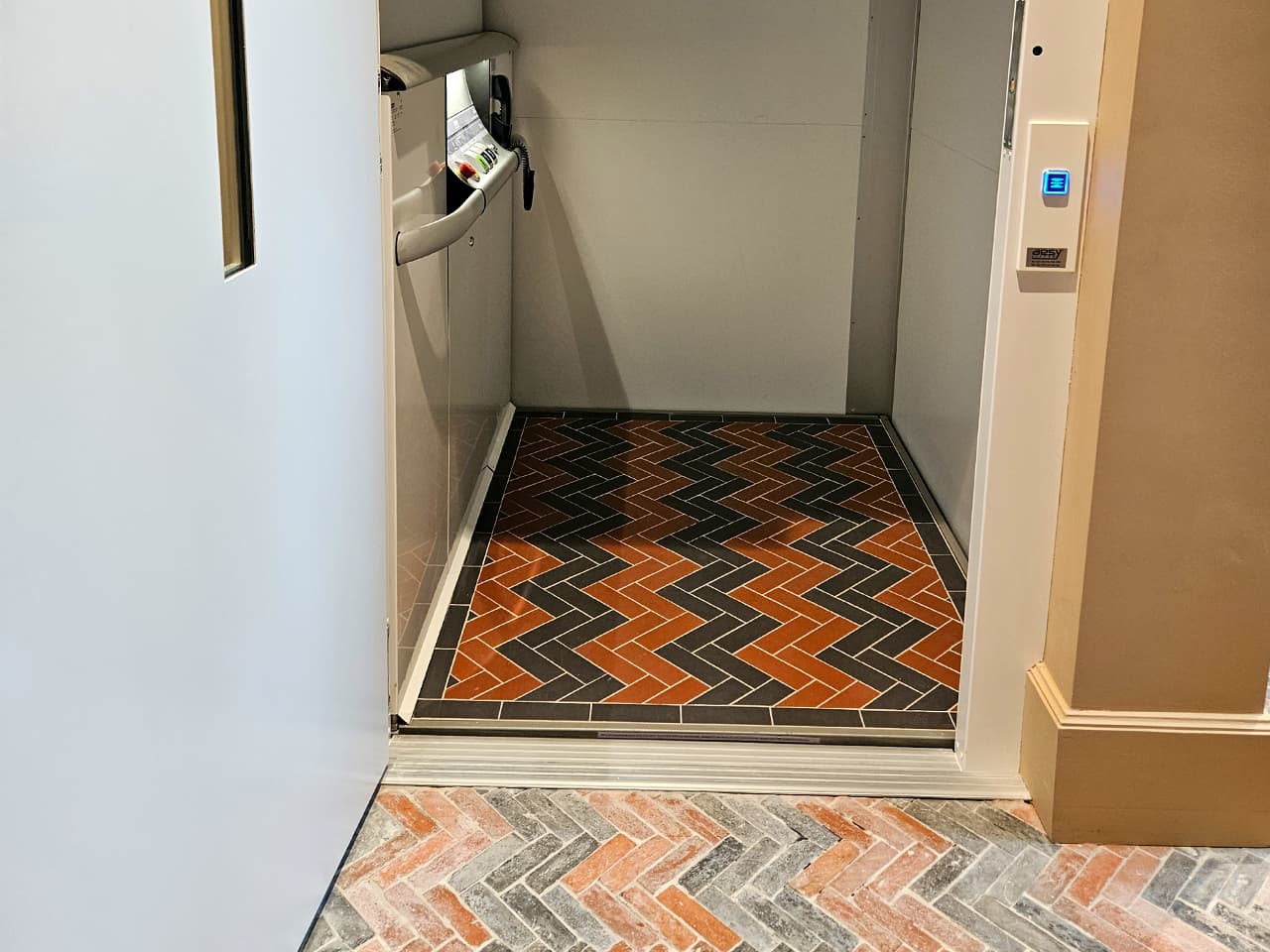 A small Cibes A5000 elevator with herringbone-patterned flooring in black, red, and gray tiles, matching the adjacent exterior floor. The elevator features a control panel inside and a call button with a blue light on the right wall.