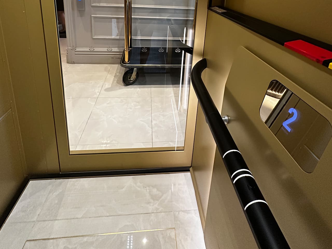 A gold-colored Cibes Air platform elevator interior with a black handrail featuring white accents on the right side. The elevator door is open, revealing a white-tiled floor and a luggage cart; a digital display shows the number "2."