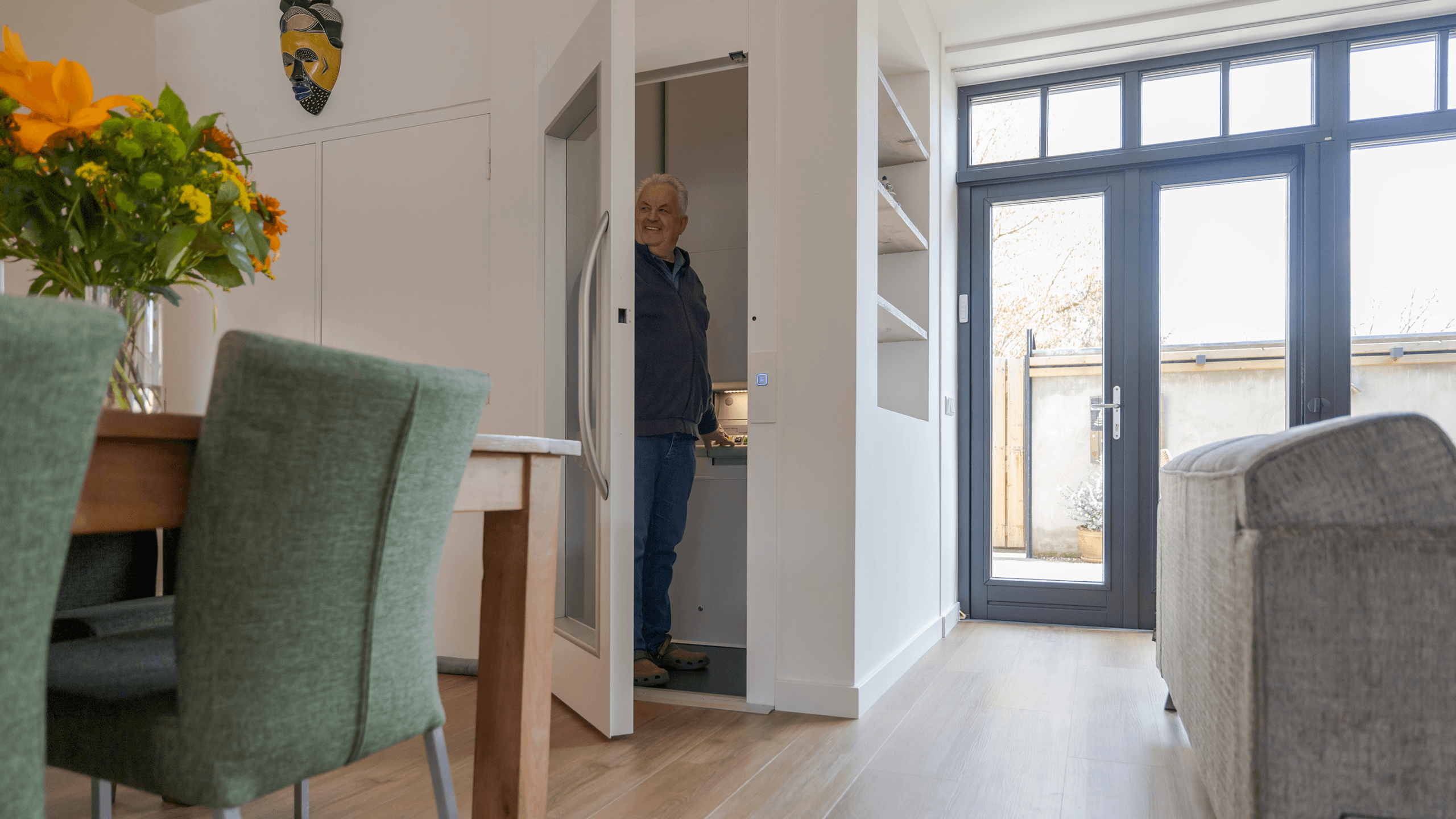 A man stands inside a compact Cibes A4000 home elevator with an open door, smiling and looking outward in a modern interior. The foreground includes a dining table with green chairs and a vase of bright flowers, while large glass doors lead to an outdoor area.