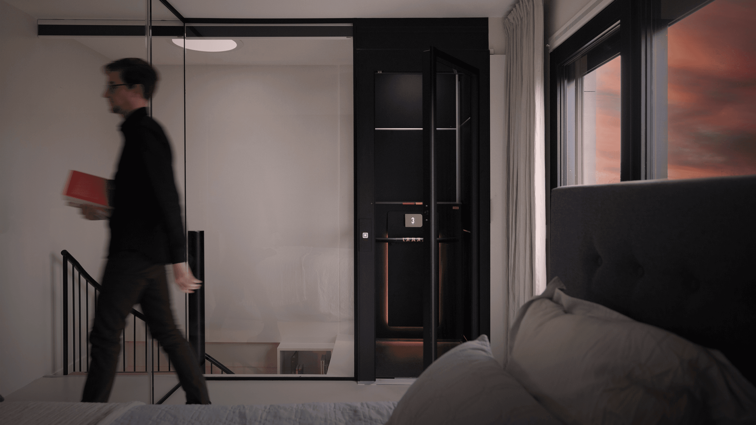 A man holding a red book walks past a modern black Cibes Air home elevator with open doors in a sleek, minimalistic interior. The foreground shows part of a bed, and large windows on the right frame a sunset sky.